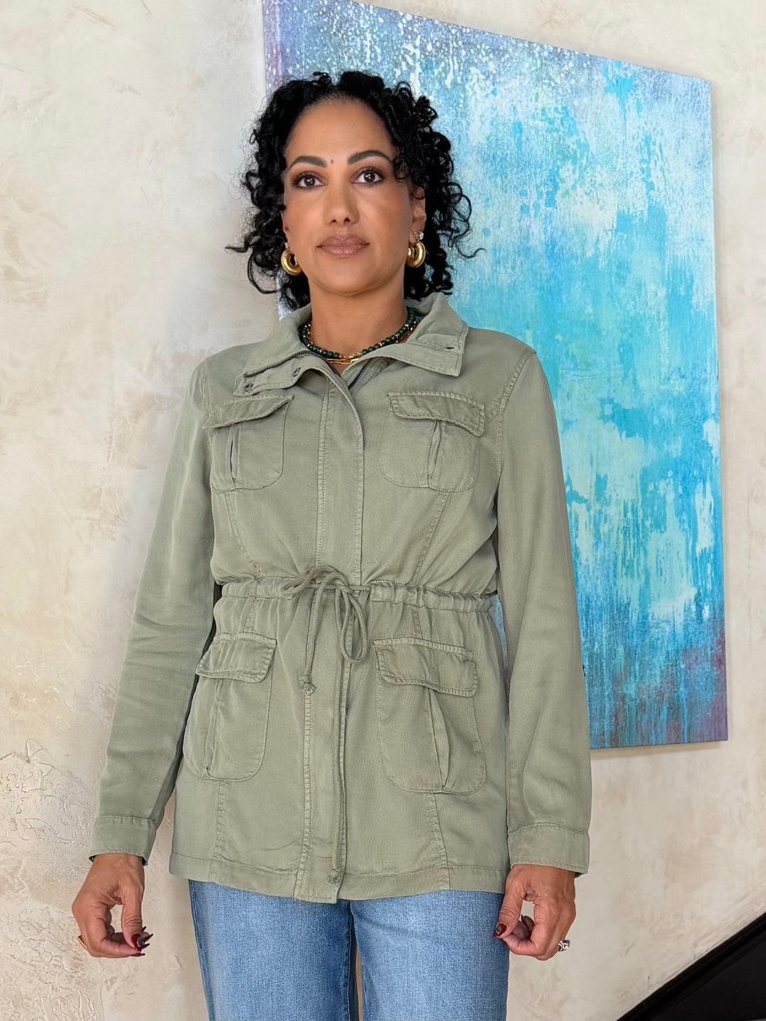 Woman wearing sage green Tencel utility jacket with drawstring waist, front zipper closure, and relaxed fit styled with wide-leg denim and neutral accessories.