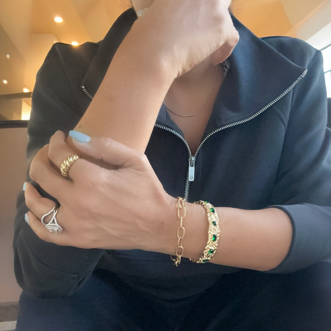 Person wearing gold rings and a bracelet with green stones indoors.