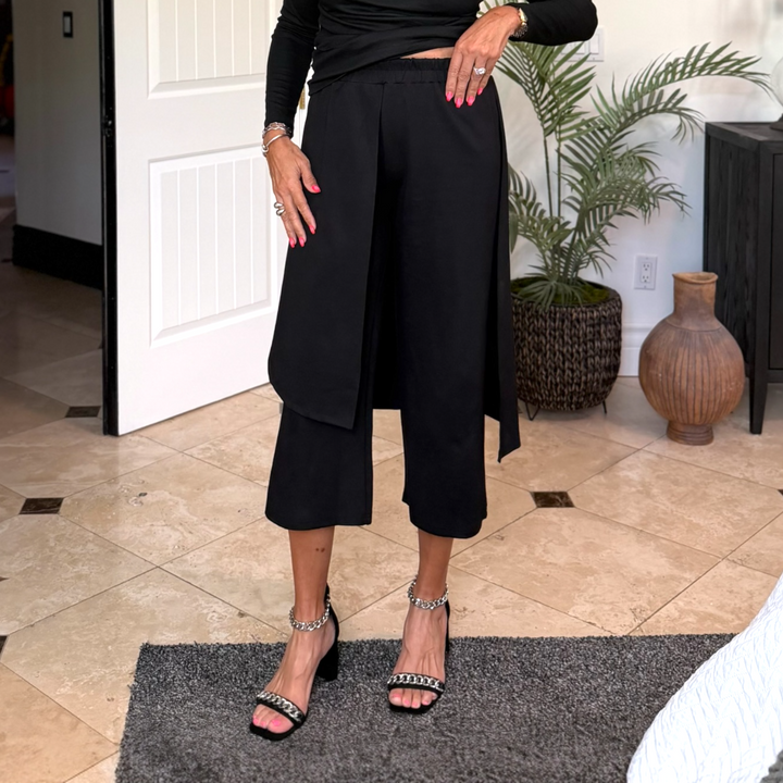 Person wearing black outfit with wide-leg pants in a room with plants and a door.