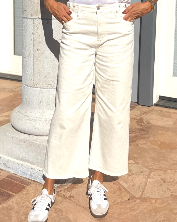 Woman wearing ivory wide-leg cropped cotton denim pants with high-rise waist, styled with white V-neck tee, brown belt, and sneakers for a modern casual outfit.