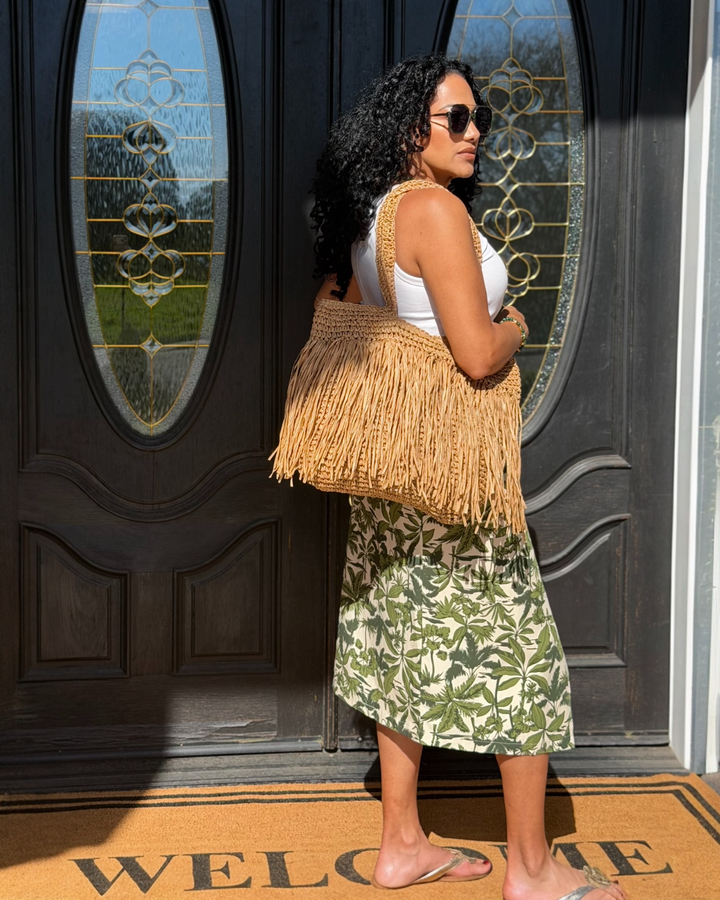 Handwoven natural raffia fringe tote bag with shoulder straps, zipper closure, and boho fringe detailing, styled with a summer outfit for a resort-inspired look.