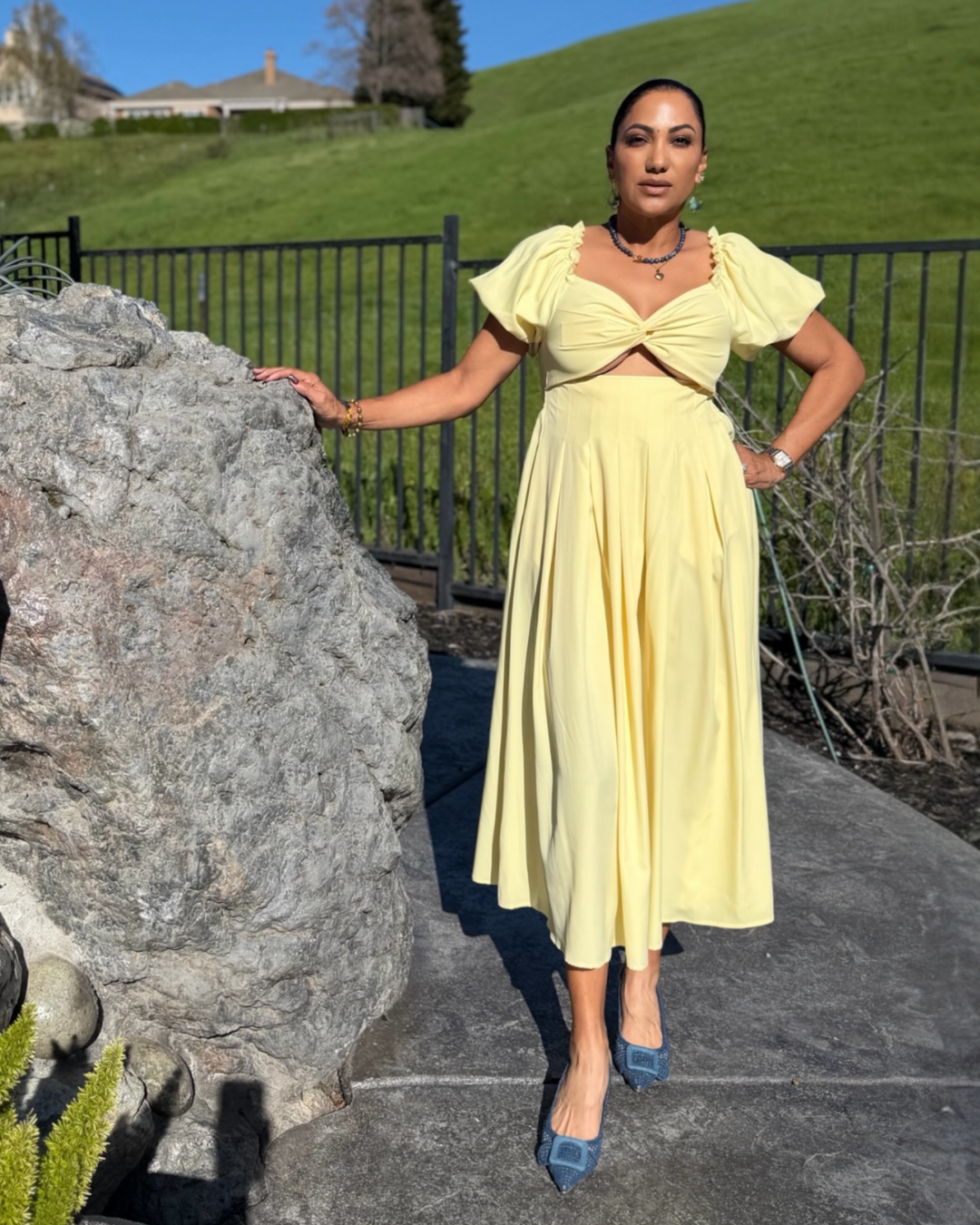 Woman wearing a soft yellow puff sleeve midi dress with a twist front bodice, styled with blue heels and a metallic clutch outdoors.