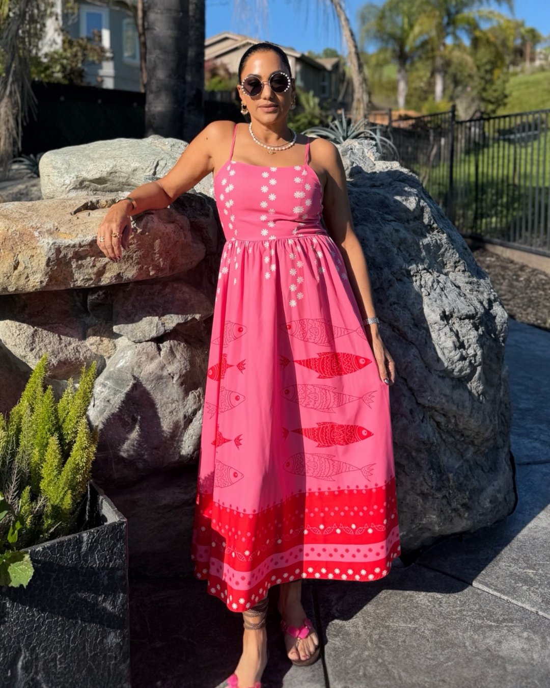 Woman wearing a pink spaghetti strap midi dress with white floral accents and red fish print along the hem, styled with sunglasses and sandals outdoors.