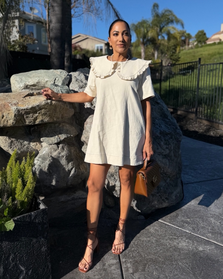Woman wearing a cream mini dress with a ruffled oversized collar, styled with lace-up heels and a brown handbag outdoors.