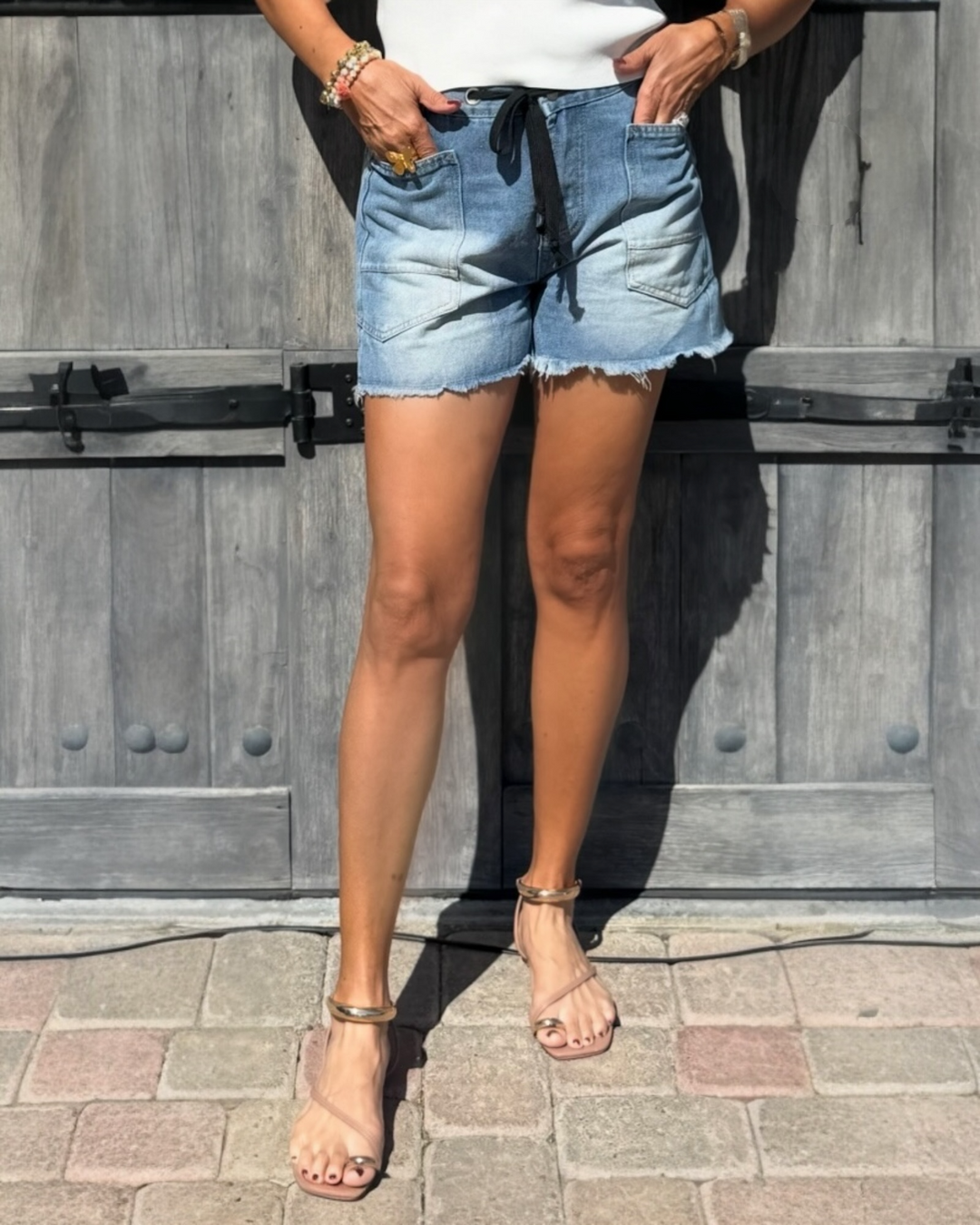 Light wash denim drawstring shorts with frayed hem styled with a white tank top and sandals, casual summer boutique outfit.