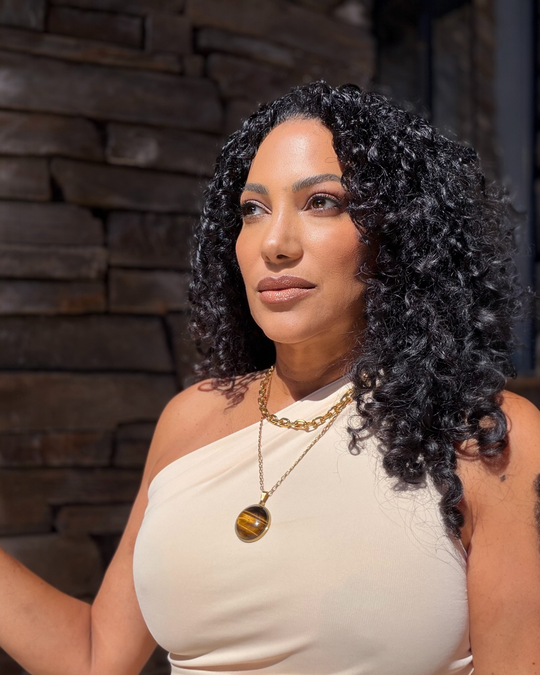 Woman with styled hair wearing a gold tigers eye necklace against a stone wall background