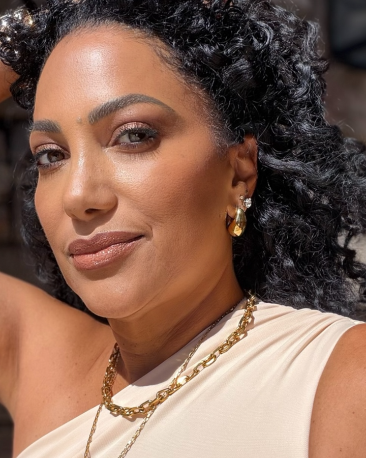 Woman with styled hair and jewelry, wearing a beige top.