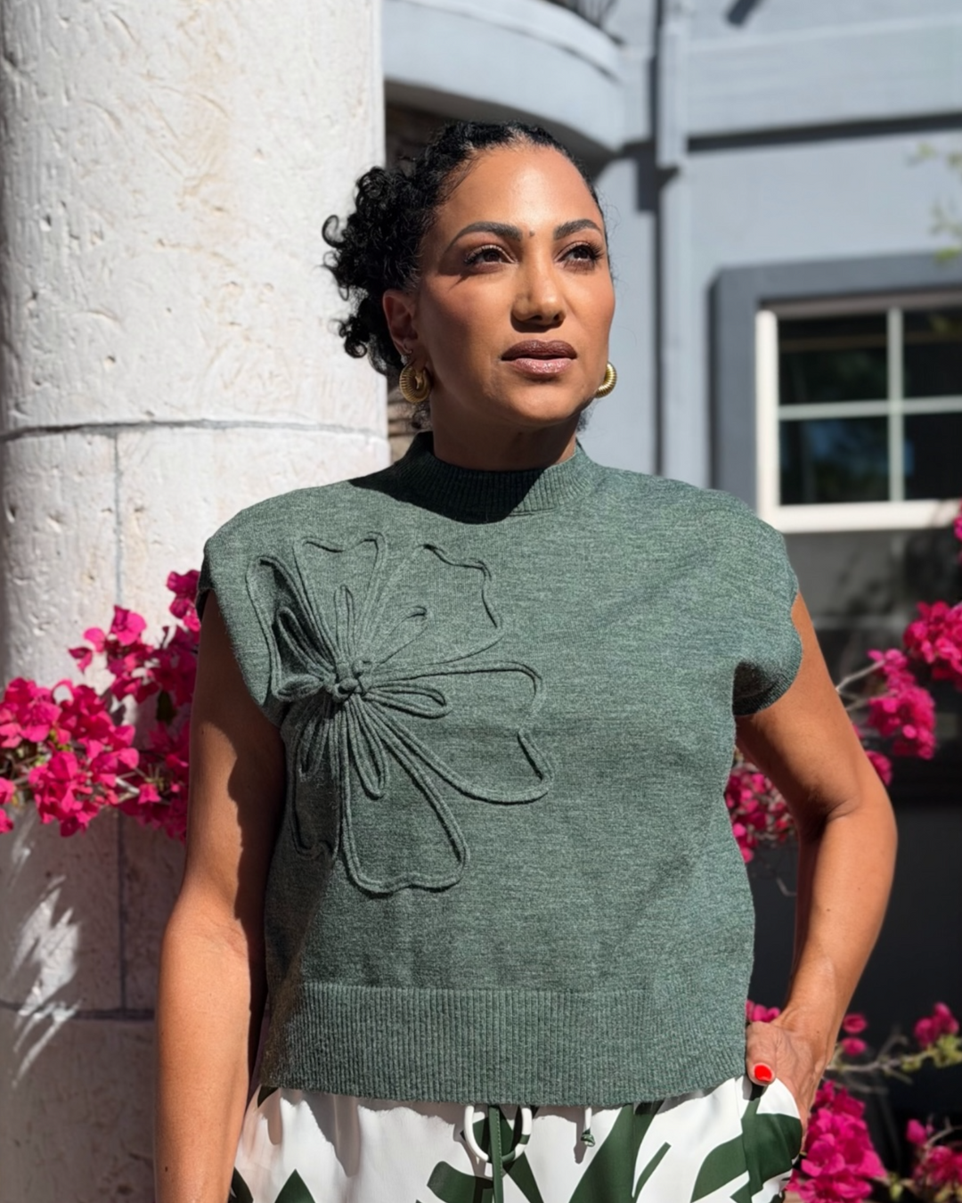 Woman wearing a green sleeveless sweater with floral details against a building and pink flowers background