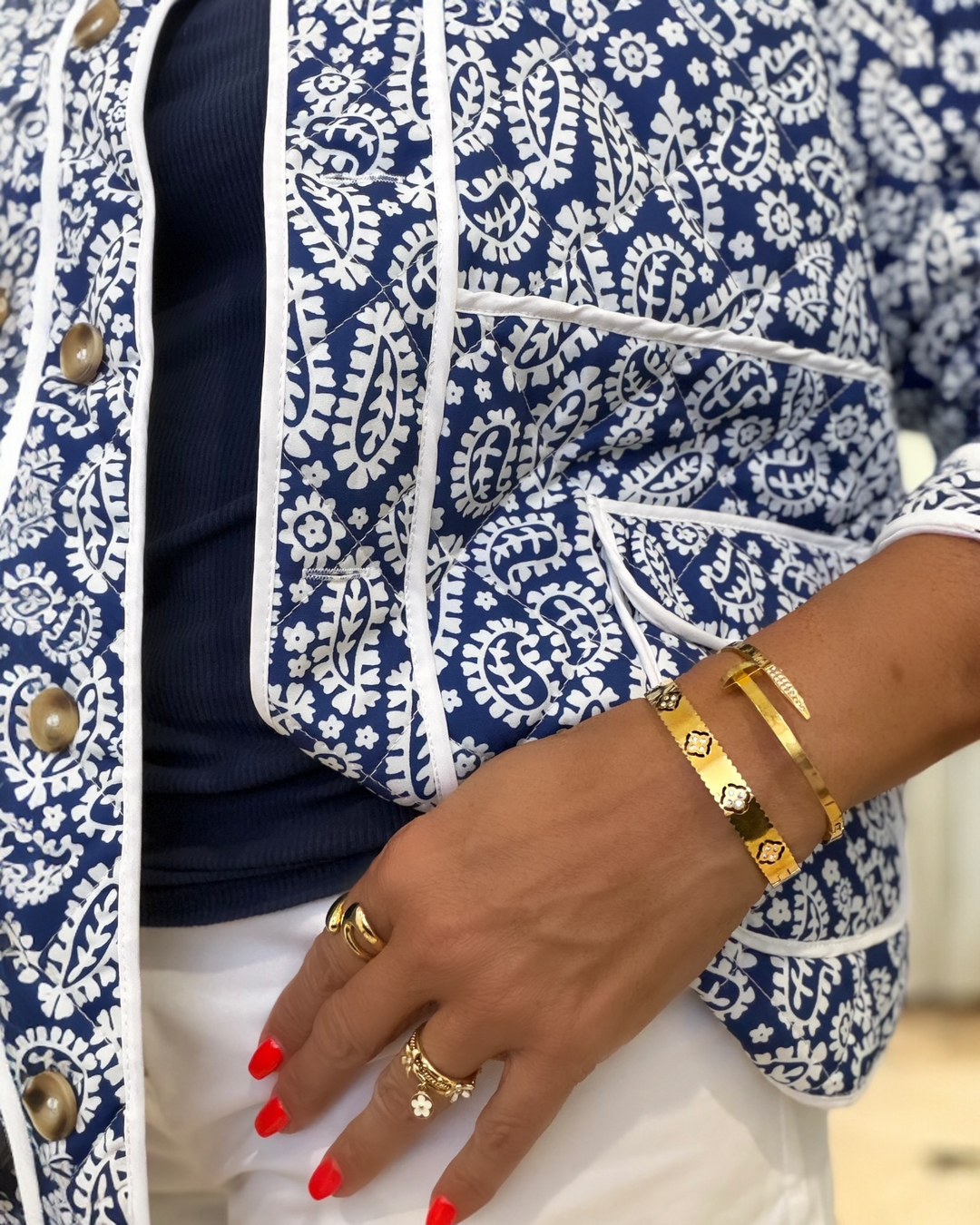Person wearing a blue and white patterned jacket with gold nail bracelet jewelry and red nail polish.
