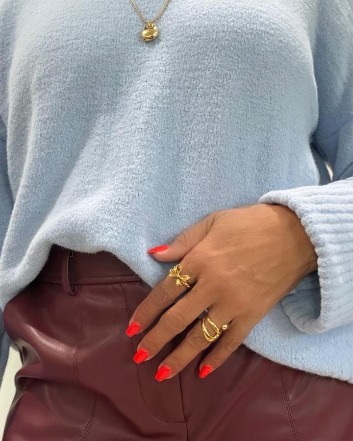 Person wearing a light blue sweater and brown pants with gold bow ring jewelry and red nail polish.