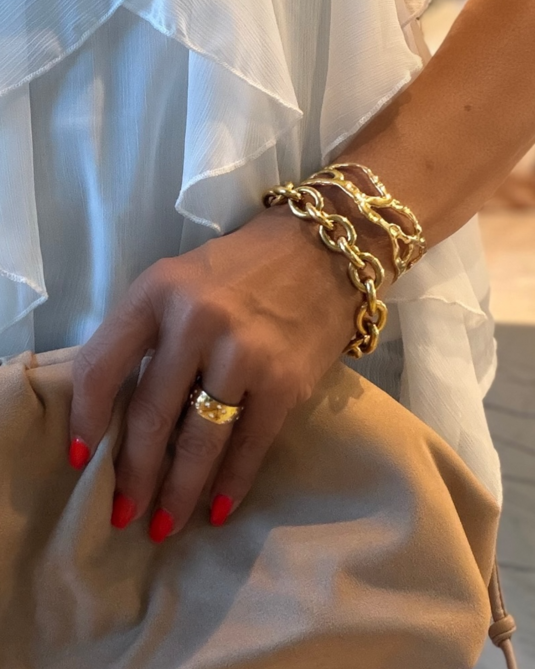 Hand wearing a gold ring and bracelet with red nail polish, against a light background