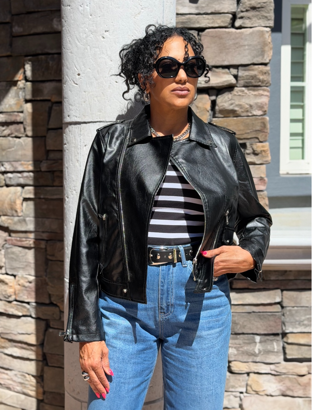 Woman wearing a black leather jacket, striped shirt, and blue jeans standing against a stone wall.
