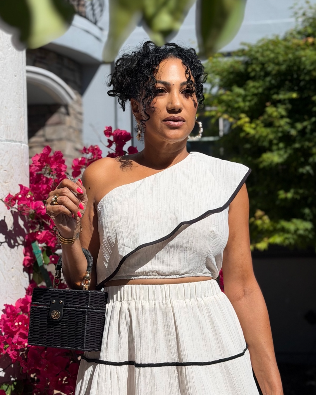 Woman wearing a cream one-shoulder textured dress with black trim detailing, styled with gold jewelry and holding a black woven handbag, standing outdoors near vibrant pink flowers in sunlight.