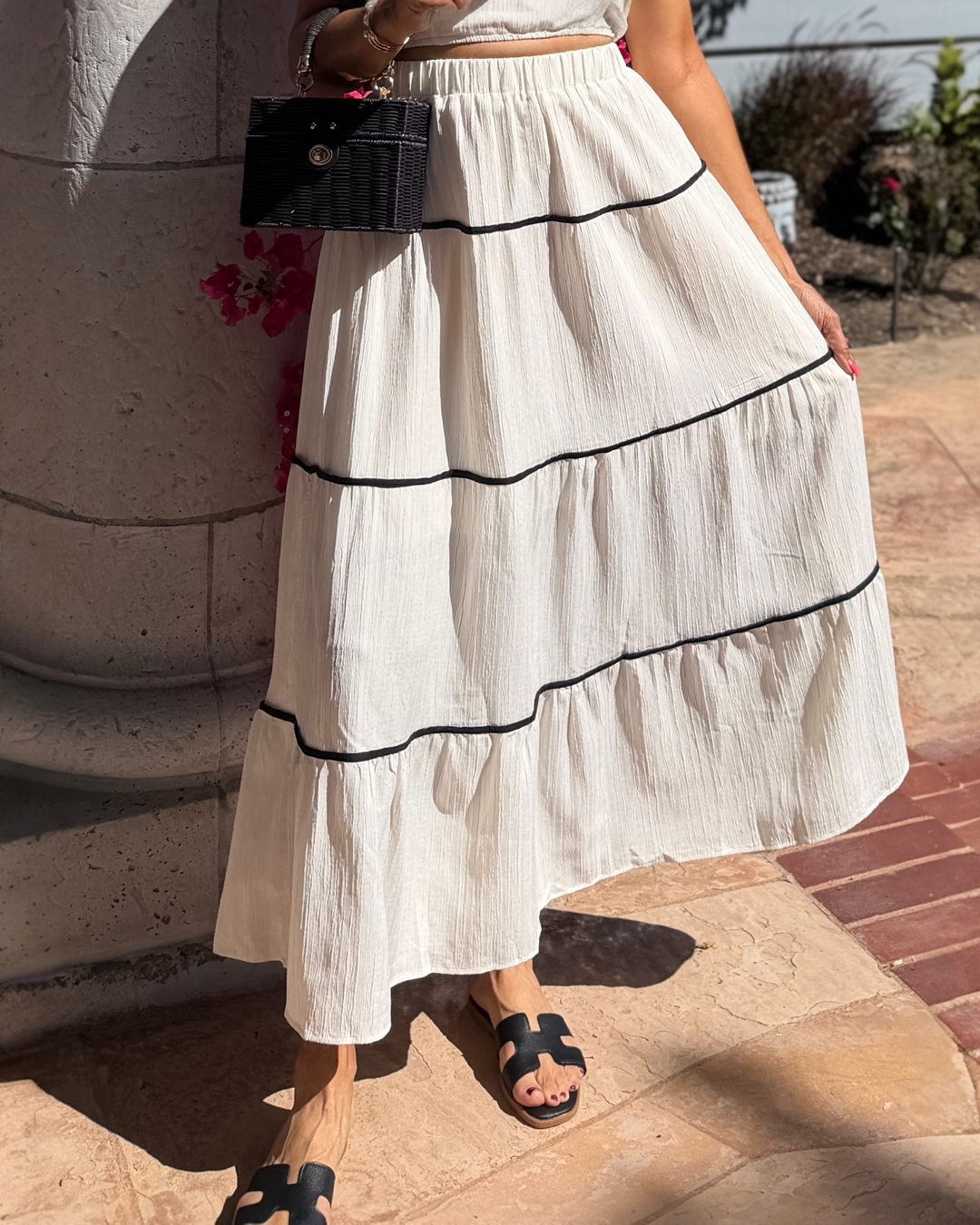 Close-up of a cream textured tiered maxi skirt with black trim detailing, styled with a matching top and black slide sandals, paired with a structured black woven handbag, photographed outdoors in sunlight.