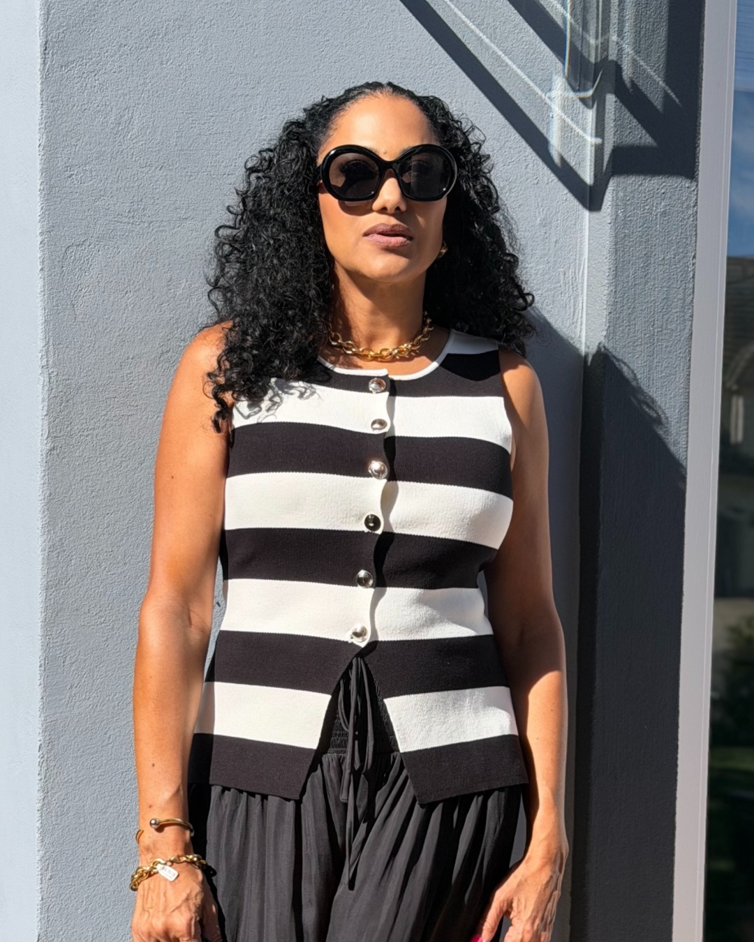 Woman wearing a black and white striped sleeveless knit top with button front detail, styled with black pants and gold accessories, standing outdoors in sunlight.
