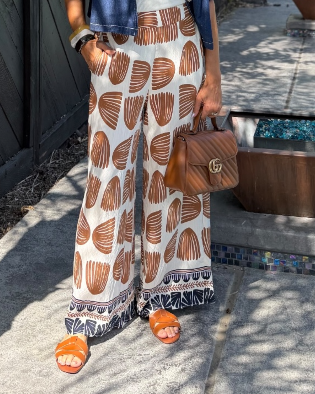 Woman modeling printed wide-leg pants in an ivory and camel abstract pattern with border hem detail, styled with a white tank, cropped denim jacket, tan handbag, and orange sandals for a chic resort-inspired look.