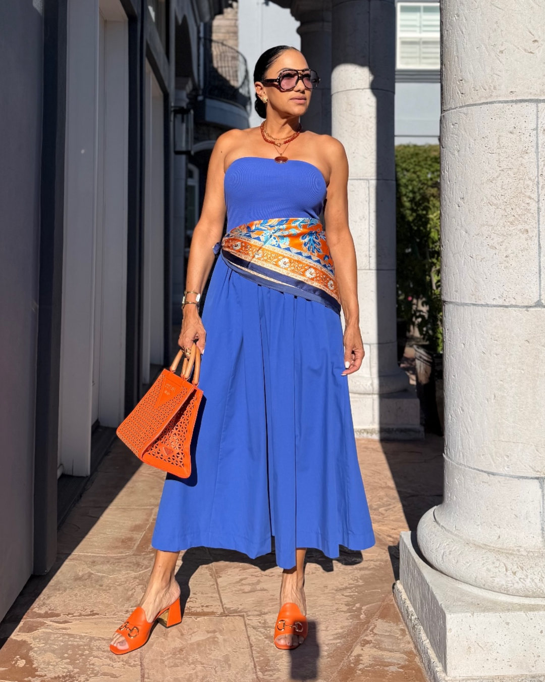 Orange and cobalt blue floral square scarf styled as a neck scarf and waist accessory with a vibrant Mediterranean-inspired print.