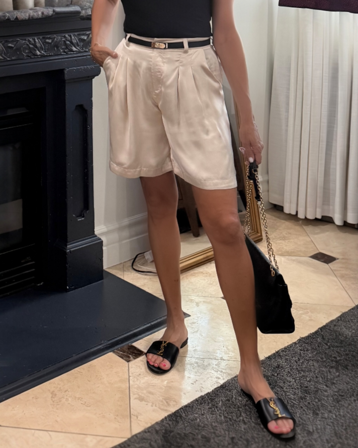 Woman wearing high-rise cream satin pleated bermuda shorts styled with a black fitted tee, slim black belt, black chain-strap bag, and black slide sandals.
