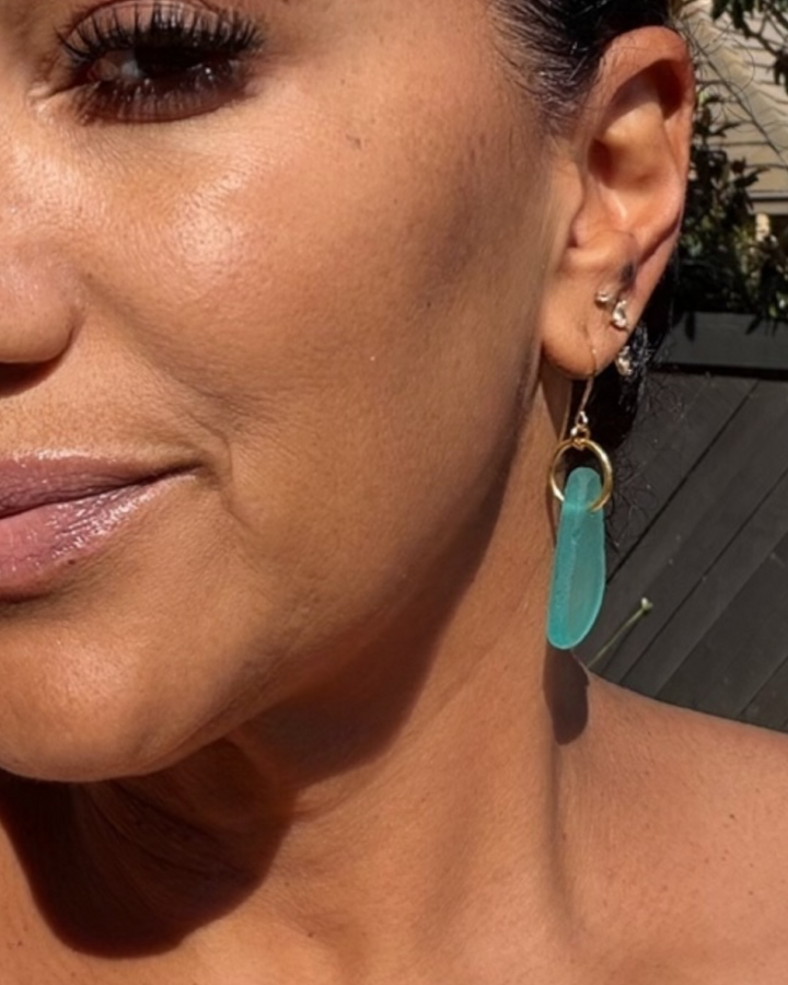 Pair of turquoise sea glass drop earrings with gold fish hook and gold ring connector, shown on model against natural light.