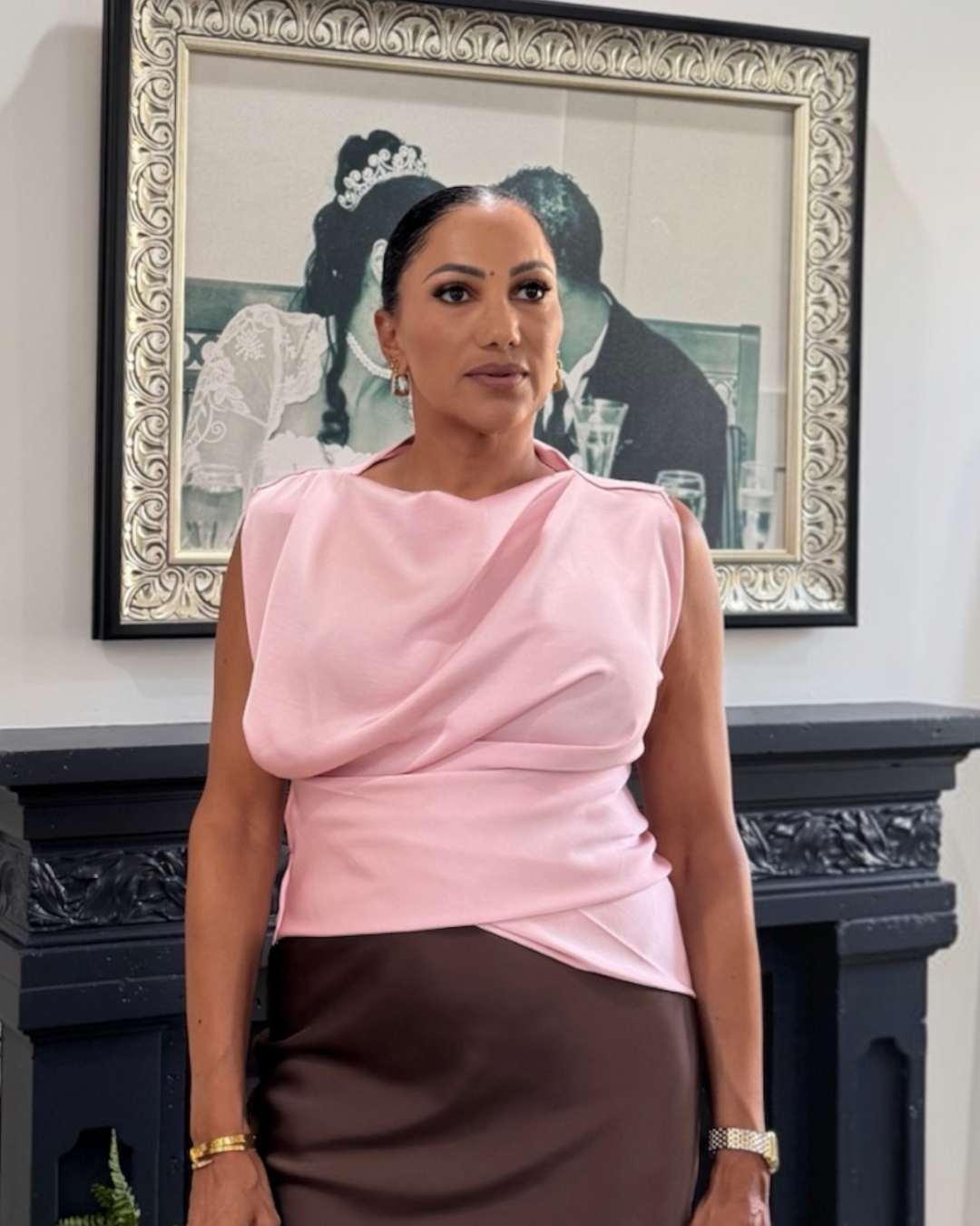 A woman wearing a light pink draped sleeveless top and a dark brown skirt, standing in front of a framed black-and-white portrait and a dark fireplace mantel.