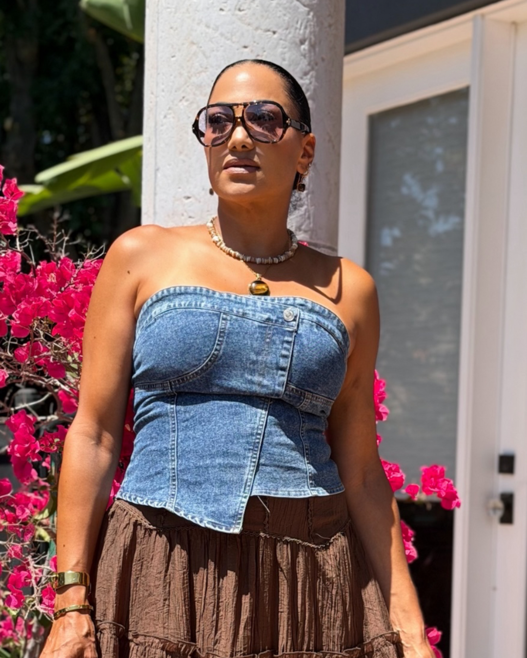 A woman wearing a blue denim strapless corset-style top and a brown pleated skirt, standing outdoors in front of a white column and door with bright pink flowers.