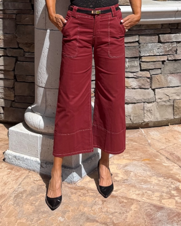 A person wearing burgundy wide-leg cropped pants with contrast stitching, paired with black pointed-toe heels, standing outdoors against a stone wall and white column.