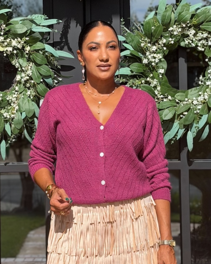 A woman wearing a magenta cable-knit V-neck cardigan with white buttons and a beige pleated skirt, standing in front of a black glass door decorated with green wreaths.