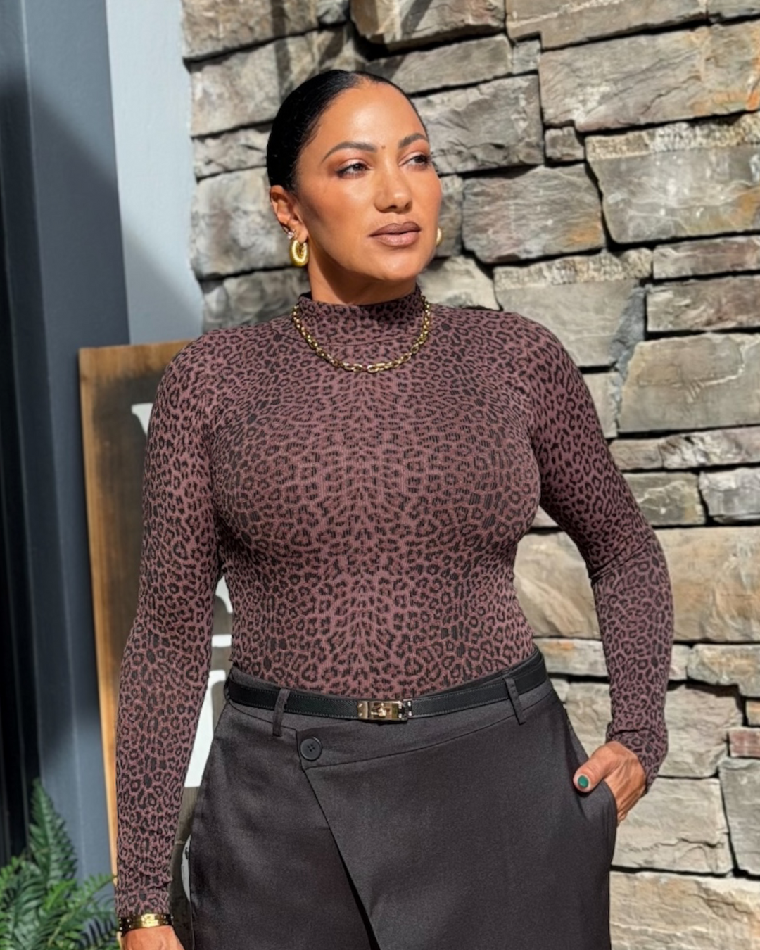 A woman wearing a fitted long-sleeve mock-neck top with a brown and black leopard print, paired with dark gray trousers and gold jewelry, standing in front of a stone wall.