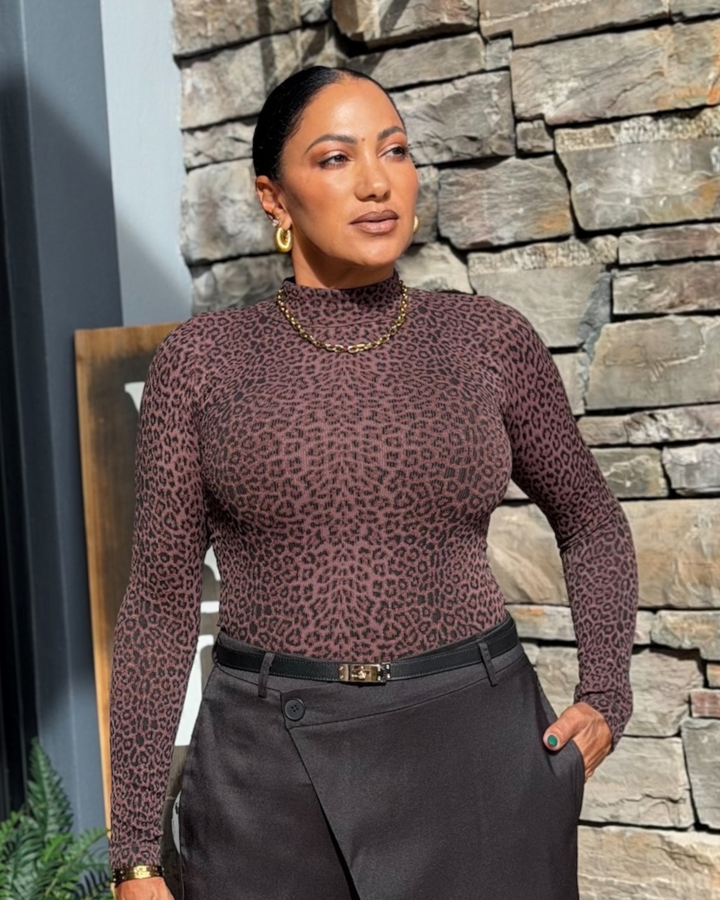 A woman wearing a fitted long-sleeve mock-neck top with a brown and black leopard print, paired with dark gray trousers and gold jewelry, standing in front of a stone wall.